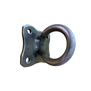 Puede incluir: Un punto de anclaje de metal gris oscuro que presenta una placa de montaje rectangular plana con dos orificios circulares para su fijación y un anillo circular grande y grueso soldado al centro, con marcas de soldadura visibles en la unión.