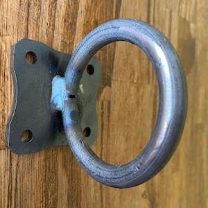 Puede incluir: Un anillo de metal negro unido a una placa cuadrada de metal con cuatro agujeros. El anillo tiene un diámetro de unos 7,5 cm y la placa tiene una anchura de unos 10 cm.