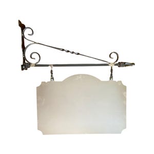 Könnte beinhalten: Ein dekorativer Schmiedeeisen-Schildhalter mit einem leeren, hellfarbigen Schild. Die Halterung weist kunstvolle Schnörkel und ein gedrehtes Metalldesign auf. Das Schild ist rechteckig mit einem dekorativen oberen Rand.