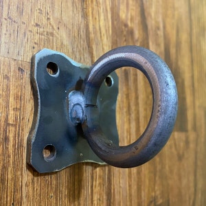 May include: A black metal ring with a square mounting plate. The ring is attached to the plate with two screws. The plate has two holes on the side.