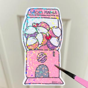 May include: A pink sticker with a gumball machine design and the text "GACHA MANIA". The machine is filled with colourful gumball-like objects. The sticker has a glittery finish.