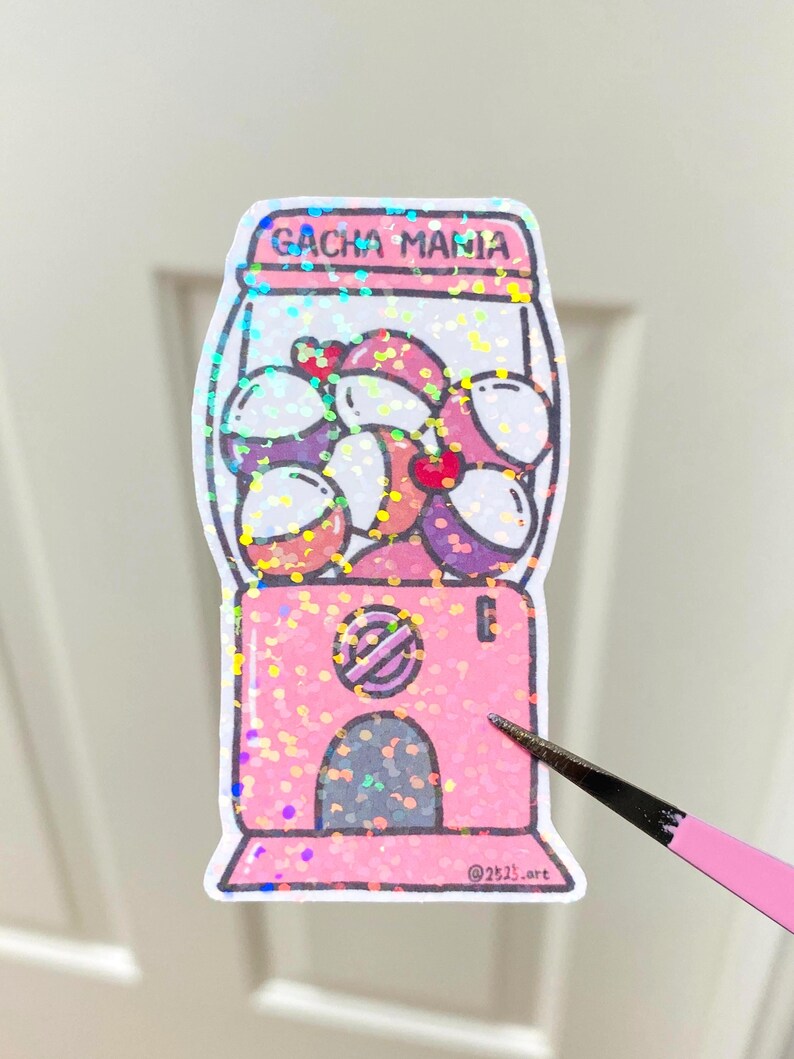 May include: A pink sticker with a gumball machine design and the text "GACHA MANIA". The machine is filled with colourful gumball-like objects. The sticker has a glittery finish.