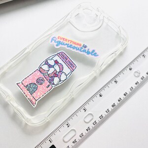 May include: Transparent phone case featuring a pink gumball machine sticker with the text "GACHA MANIA". The case also has a sticker that says "Everything is Figureoutable". A clear ruler is next to the phone case.