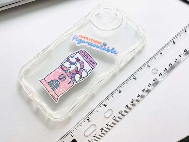 May include: Transparent phone case featuring a pink gumball machine sticker with the text "GACHA MANIA". The case also has a sticker that says "Everything is Figureoutable". A clear ruler is next to the phone case.