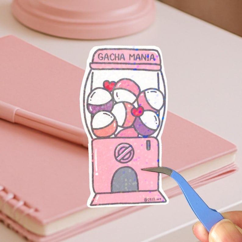 May include: A pink sticker depicting a gumball machine with the words "GACHA MANIA". The machine contains colourful gumball-like objects. A pair of blue tweezers is holding the sticker.