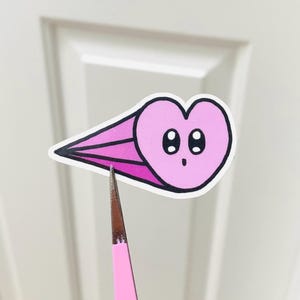 May include: A pink heart-shaped sticker with a surprised expression and black outlines. The sticker has a comet-like design with pink and purple streaks. Held by pink tweezers.