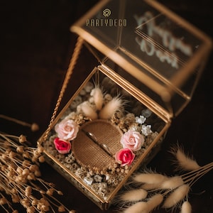 Puede incluir: Una caja de vidrio con un borde dorado y una tapa con cadena, llena de flores secas y dos anillos sobre un cojín de arpillera. La tapa tiene el texto "Partydeco".