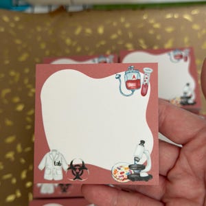 May include: A stack of square sticky notes with a light red border and a white center. Each note features watercolor illustrations of lab equipment, including a microscope, blood bag, and lab coat. The word "lab" is printed on the lab coat.
