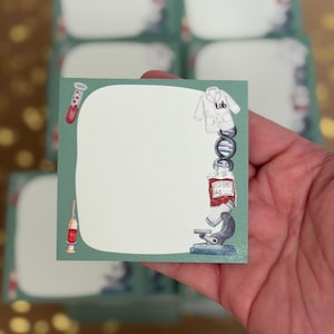 May include: A square, green sticky note with a white center and science-themed illustrations. The illustrations include a lab coat, DNA strand, blood bag, microscope, and test tube. The word "Lab" is printed on the lab coat.