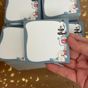 May include: A white ceramic mug with a black handle and the text "But First, Coffee" in a black, handwritten-style font. The mug sits on a white surface.