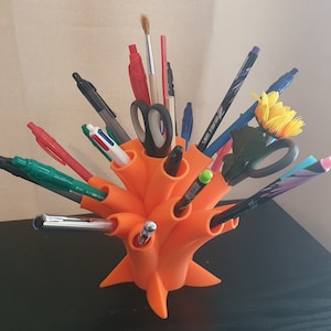 May include: Orange 3D printed desk organizer with multiple slots for pens, pencils, and other office supplies. The organizer is filled with various writing utensils, including a pair of scissors, a paintbrush, and a small artificial sunflower.