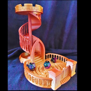 Puede incluir: Una torre dorada impresa en 3D con una escalera de caracol y una plataforma circular en la base. La torre tiene una pequeña puerta arqueada y una barandilla decorativa. Hay tres dados en la plataforma, dos de los cuales son multicolores.