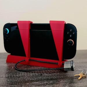 以下が含まれることがあります： 黒い携帯型ゲーム機が赤いプラスチックホルダーに固定されています。黒いケーブルと鍵付きの銀色の南京錠がホルダーに取り付けられています。デバイスには青とオレンジのボタンがあります。