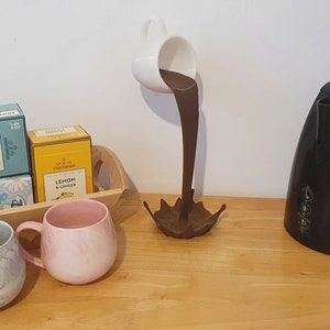 May include: A wooden tray with two boxes of tea, one labeled "Peppermint" and the other "Lemon & Ginger". There are two mugs, one gray and one pink, and a black electric kettle. A white ceramic mug is pouring brown liquid into a metal sculpture of a splash.