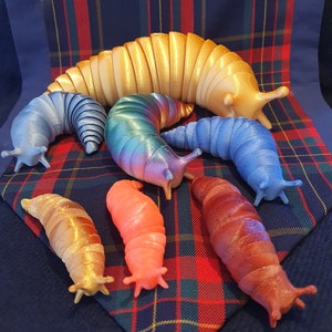 Op de afbeelding: Een verzameling van zeven kleurrijke, gesegmenteerde, slak-achtige figuurtjes. De slakken zijn gemaakt van een glad, glanzend materiaal en zijn gerangschikt op een rood-zwarte geruite stof.