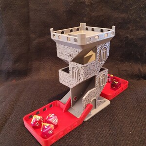 May include: A 3D printed grey tower-shaped dice rolling game with a red tray. The tower has a stone-like texture and the tray has a raised edge. Three multi-coloured dice with white numbers are in the tray.