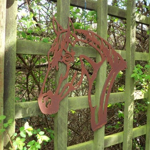 Puede incluir: Una silueta de cabeza de caballo de metal oxidado montada en una valla de madera. La cabeza de caballo es una pieza de arte decorativa para el jard&iacute;n.