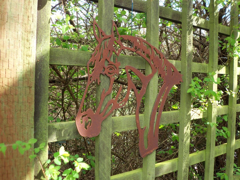 Puede incluir: Una silueta de cabeza de caballo de metal oxidado montada en una valla de madera. La cabeza de caballo es una pieza de arte decorativa para el jard&iacute;n.