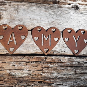 May include: Three rusty metal hearts with the name "AMY" spelled out in the center of each heart. The hearts are arranged in a row on a wooden surface.