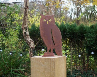 May include: A flat, rust-coloured metal owl silhouette sculpture featuring cut-out eye details. The owl figure is mounted upright on a square, light-coloured wooden post, displaying a weathered, oxidised finish across its entire surface.