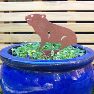 May include: A rust-colored metal garden stake shaped like a capybara silhouette, featuring a textured, weathered finish. The animal figure is positioned upright within a vibrant, deep blue ceramic planter filled with small green leafy plants.