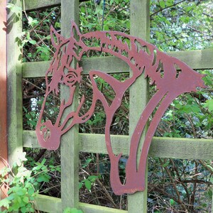 Puede incluir: Una silueta de cabeza de caballo de metal oxidado montada en una valla de madera. La cabeza de caballo est&aacute; recortada en metal y tiene un dise&ntilde;o detallado. La valla es de madera y tiene una p&aacute;tina verde.