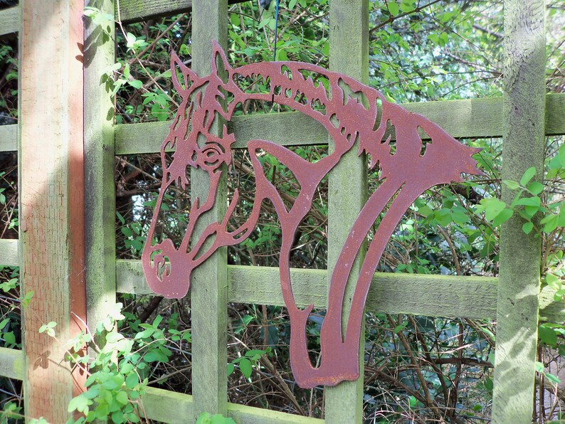 Puede incluir: Una silueta de cabeza de caballo de metal oxidado montada en una valla de madera. La cabeza de caballo est&aacute; recortada en metal y tiene un dise&ntilde;o detallado. La valla es de madera y tiene una p&aacute;tina verde.