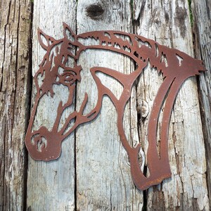 Puede incluir: Una silueta de cabeza de caballo de metal oxidado, montada sobre una superficie de madera desgastada. La cabeza del caballo est&aacute; mirando hacia el lado izquierdo de la imagen.