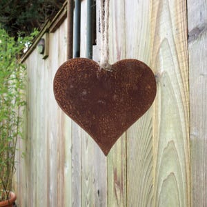 May include: A heart-shaped, rusty metal ornament hangs from a twine cord. The brown heart is suspended against a wooden fence. The image suggests a garden decoration or outdoor feature.