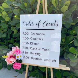 May include: A white sign on a wooden easel displays the text "Order of Events" in elegant script, followed by a schedule. A bouquet of pink and white flowers adorns the base. The background is lush green foliage.