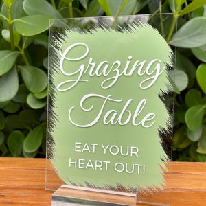 May include: A clear acrylic sign with a green painted background and white text that reads "Grazing Table Eat Your Heart Out!"