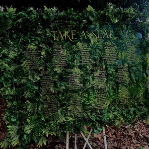May include: A clear acrylic seating chart with gold lettering that reads "TAKE A SEAT". The chart is surrounded by lush green foliage and lists names under table numbers. The chart is supported by a white metal stand.