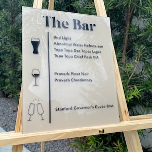 May include: A white sign with black text on a wooden easel. The sign reads "The Bar" and lists the drinks available: Bud Light, Abnormal Weiss Hefeweizen, Topa Topa Dos Topas Lager, Topa Topa Chief Peak IPA, Proverb Pinot Noir, Proverb Chardonnay, and Stanford Governor's Cuvée Brut.