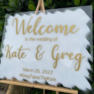 May include: White acrylic wedding welcome sign with gold lettering. The sign reads "Welcome to the wedding of Kate & Greg, March 26, 2022, #GregTakesTheKate".