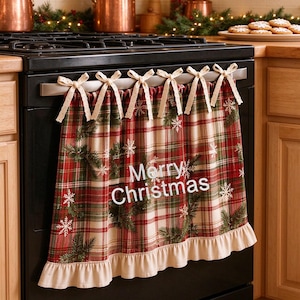 Puede incluir: Un paño de horno a cuadros rojos, verdes y crema colgado del asa de una estufa negra. El paño tiene un borde inferior con volantes, patrones de copos de nieve blancos y el texto Merry Christmas en blanco, sujeto con seis lazos de tela crema.