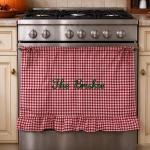 May include: A red and white gingham oven cover featuring a ruffled bottom edge and tie-top fasteners. The fabric is embroidered with the dark green text "The Briskie" centred on the front, hanging over the door of a stainless steel stove.