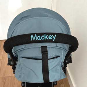 May include: A blue baby stroller with a black handle. The word "Mackey" is embroidered in blue on the handle. The stroller has a black frame and black wheels. The stroller is in a room with a wooden floor.