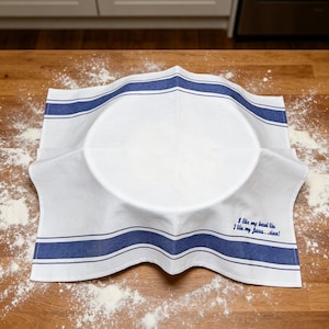 May include: A white kitchen towel with blue stripes draped over a bowl. The towel features embroidered blue text that reads, I like my bread like I like my Jesus... risen! The fabric has a smooth texture and is positioned in a square shape.