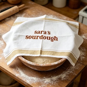 Peut inclure: Un torchon de cuisine blanc et beige rayé avec le texte "sara's sourdough" en lettres marron, couvrant un bol en bois de pâte. La scène se déroule sur une table en bois avec de la farine et des ustensiles de boulangerie.