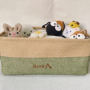 Peut inclure: Un panier de rangement rectangulaire avec un dessus beige et un fond vert, contenant plusieurs petits jouets crochetés à la main. Les jouets comprennent un lapin, une créature blanche, une marguerite et d'autres figurines d'animaux. Le mot "Rocky" est imprimé sur le devant.