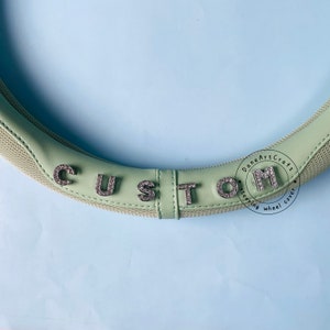 Peut inclure: Un couvre-volant de couleur vert clair avec le mot "CUSTOM" &eacute;crit en strass. Le couvre-volant a un design en maille et les mots "DaneArtCraft steering wheel cover" sont imprim&eacute;s sur le c&ocirc;t&eacute;.