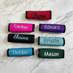 May include: A collection of colorful luggage handle wraps, each embroidered with a different name. The wraps are in various colors, including red, pink, purple, black, teal, blue, and green. The names are embroidered in white thread.