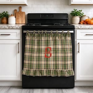 May include: A green and beige plaid fabric stove cover featuring a ruffled bottom edge and a red embroidered letter S monogram in the center. The cover is attached to the oven handle with fabric ties along the top edge of the textile panel.