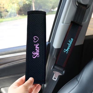 May include: Black seat belt covers with embroidered names. One cover has "Sheri" in pink script with a heart, the other has "Freddie" in blue script. Both covers are in a car interior.