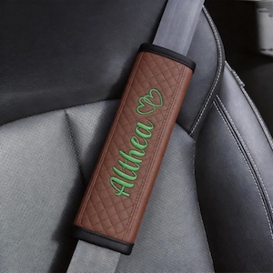 May include: A brown, quilted seat belt cover featuring the name Althea embroidered in green script, accompanied by two small green heart outlines positioned above the text. The cover has black trim and is attached to a gray seat belt strap.