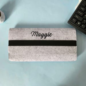 Könnte beinhalten: Hellgraues Filzetui mit dem Namen "Maggie" in schwarzer Schrift. Ein schwarzes Gummiband umschließt die Mitte. Das Etui ist rechteckig und scheint geschlossen zu sein. Der Hintergrund ist eine hellblaue Oberfläche.