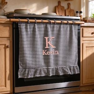 May include: A black and white gingham kitchen towel hanging from a wooden rod on an oven door. The towel features a ruffled bottom edge and is embroidered with a large pink letter K above the name Kevin in a matching pink script font.