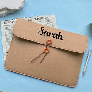 Op de afbeelding: Een beige vilten laptoptas met de naam "Sarah" in zwart script. De tas heeft een klepsluiting met twee houten knopen en touwtjes. Een pen ligt op een lichtblauwe ondergrond.