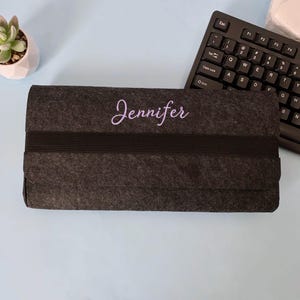 Könnte beinhalten: Eine dunkelgraue Filztasche mit dem Namen "Jennifer" in Schreibschrift gestickt. Ein schwarzes Gummiband umhüllt die Mitte. Eine Tastatur und eine kleine Sukkulente sind im Hintergrund zu sehen.