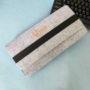May include: A light gray felt laptop sleeve with the name "Alisa" embroidered in orange. A black elastic band secures the sleeve closed. A keyboard is visible in the background.